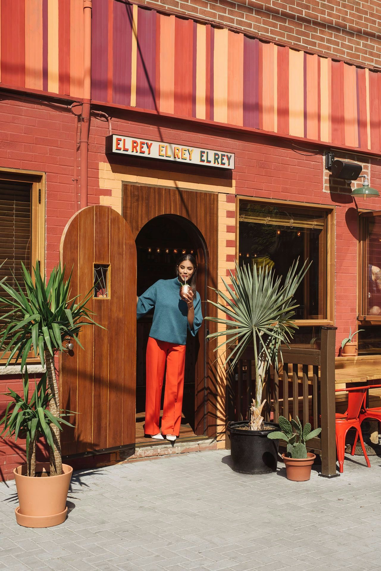A cheerful woman stands in a doorway sipping a beverage, framed by the wooden entrance of Toronto's "EL REY" restaurant with a warm, inviting exterior and potted plants