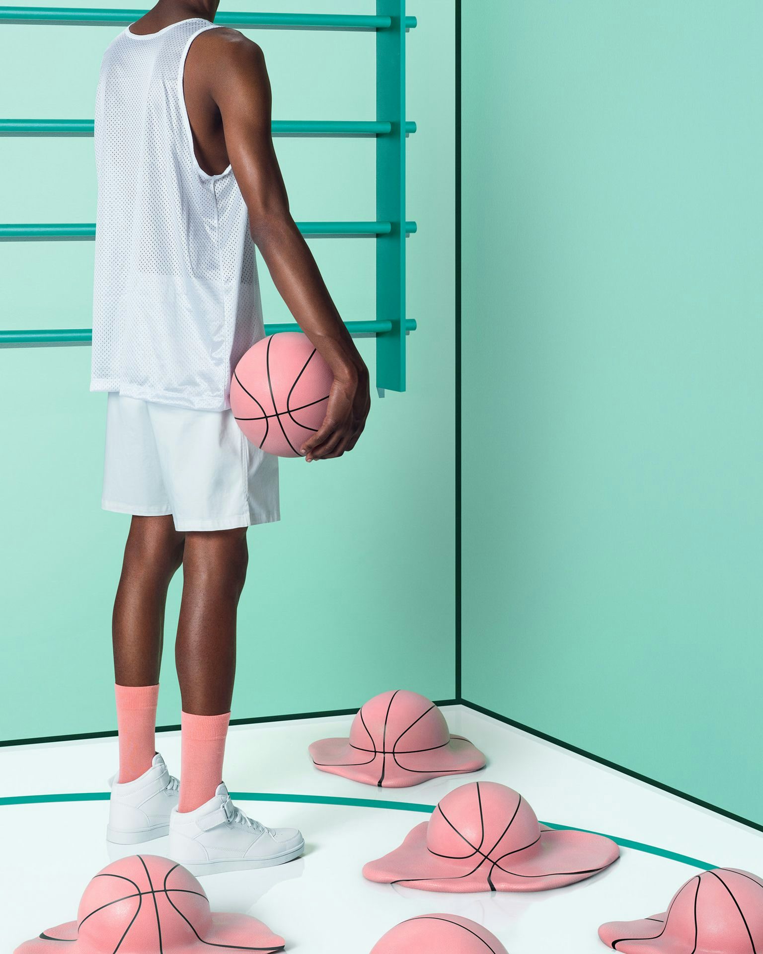 A basketball player stands, holding a ball, amidst deflated pink basketballs on a turquoise floor with a matching backdrop.