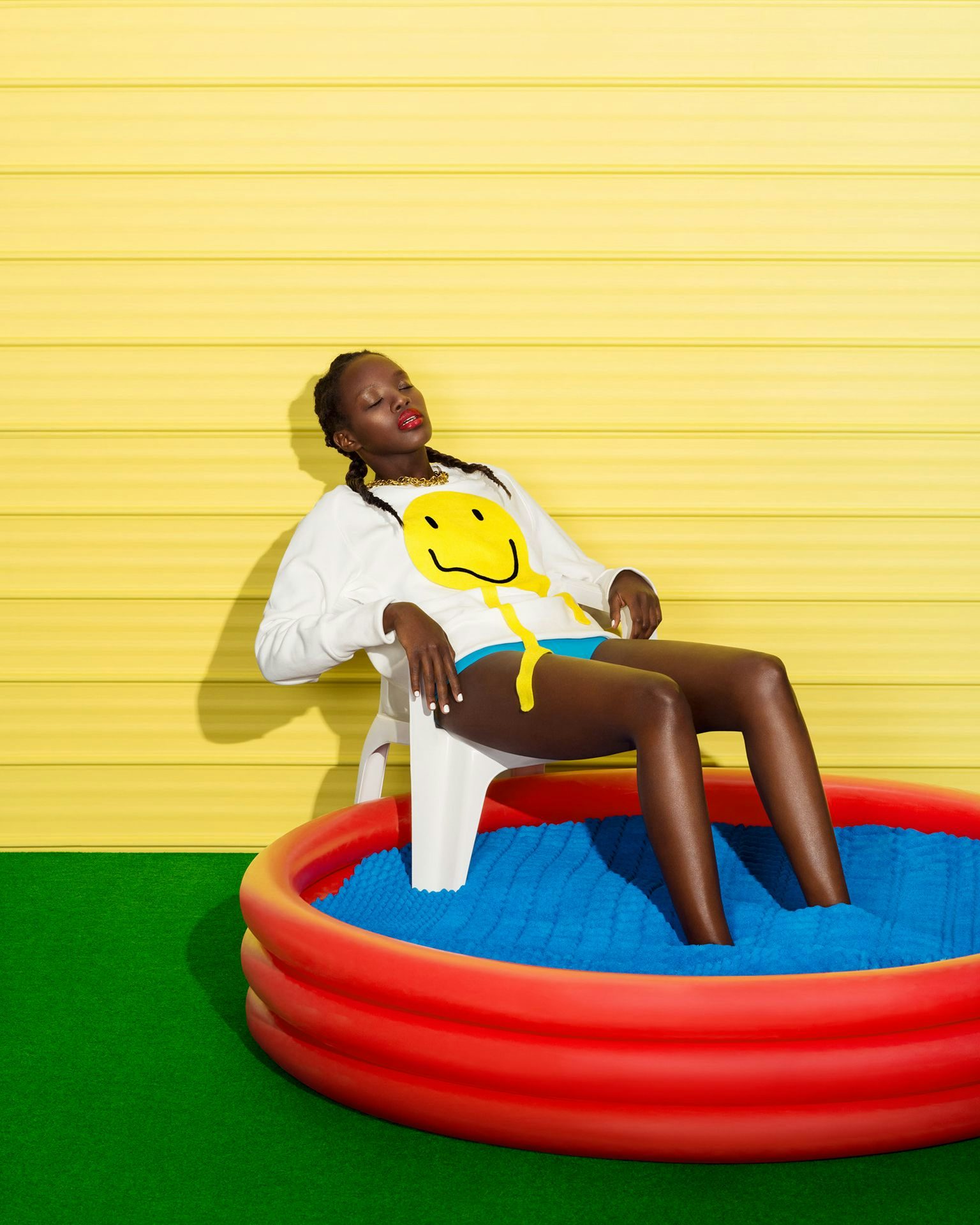 A woman relaxes in a red kiddie pool filled with blue balls, against a vibrant yellow corrugated wall backdrop, wearing a smiley face shirt.