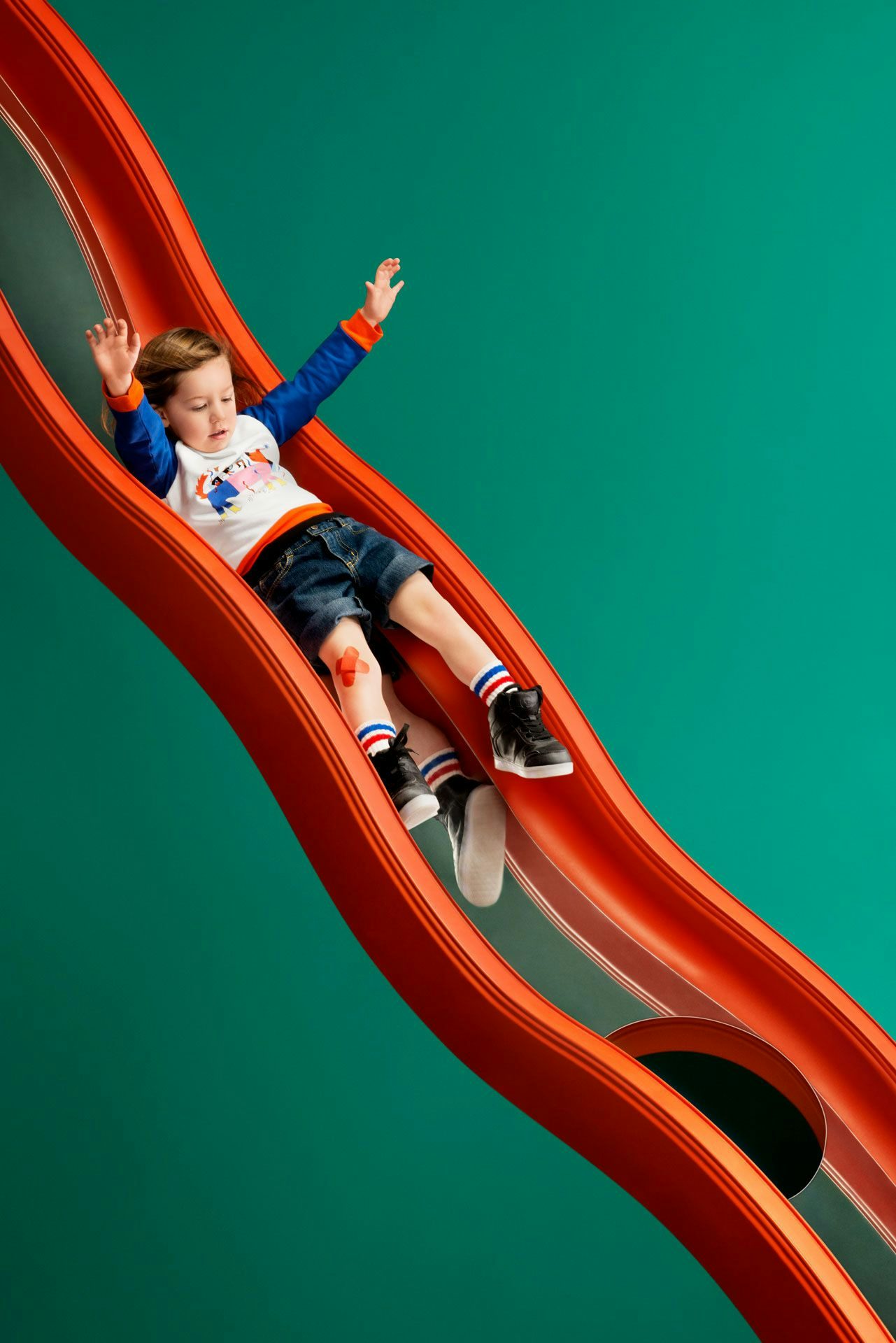A child joyfully sliding down a vibrant red slide against a teal background.