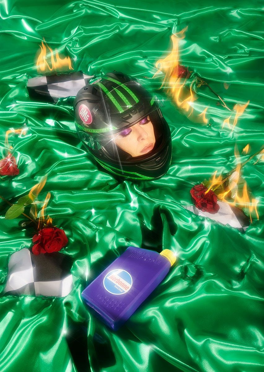 A surreal image of a woman's head in a racing helmet, lying on a green satiny fabric with flames, roses, and a bottle of motor oil.