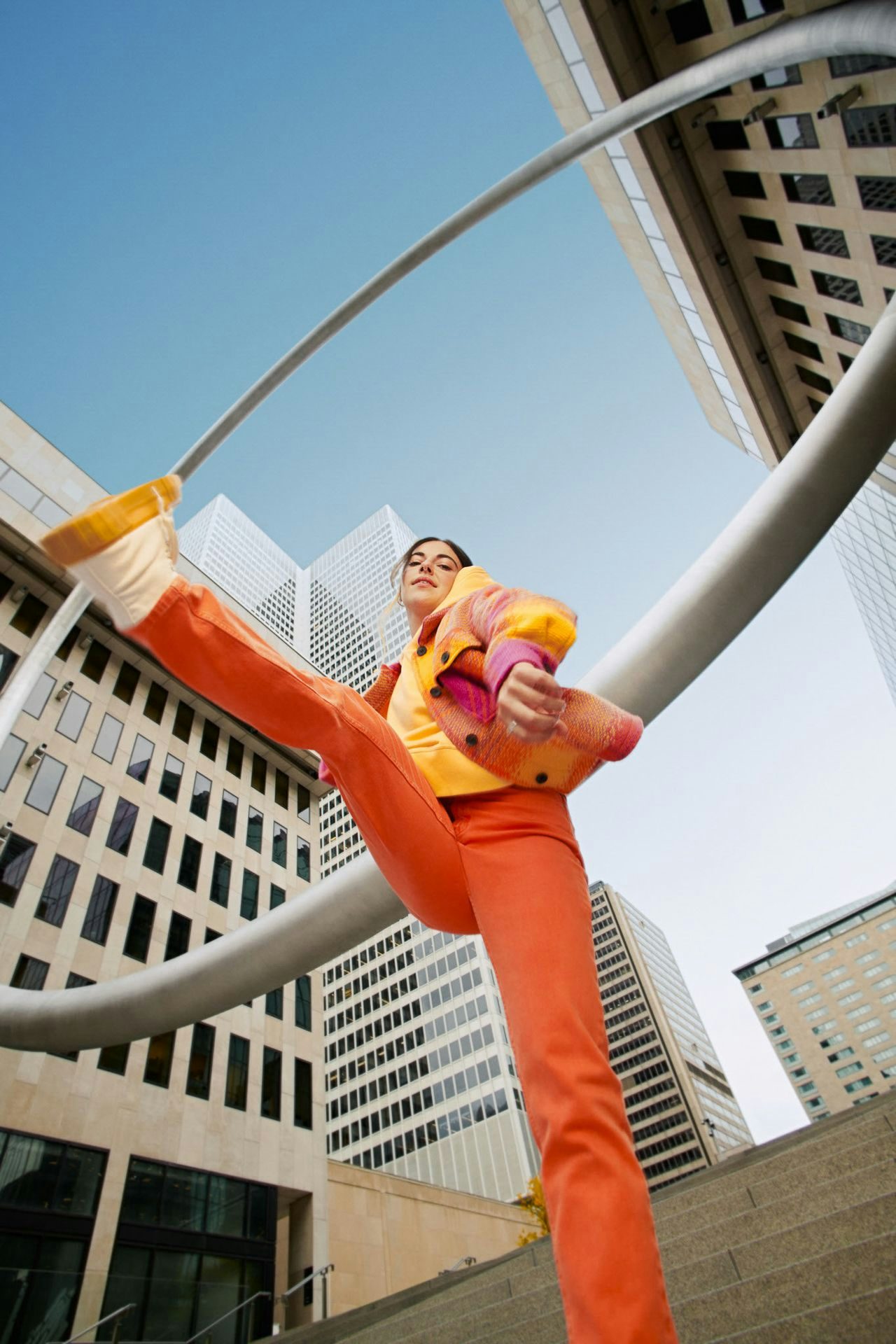  A dynamic upward angle of a person in vibrant orange pants and a colorful jacket posed against the backdrop of L'Anneau in downtown Montreal.