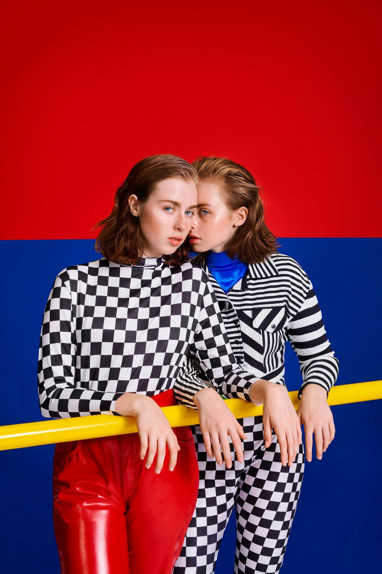 Two identical twins in checkered outfits against a red and blue background, one in a contemplative pose and the other with a whispering gesture.