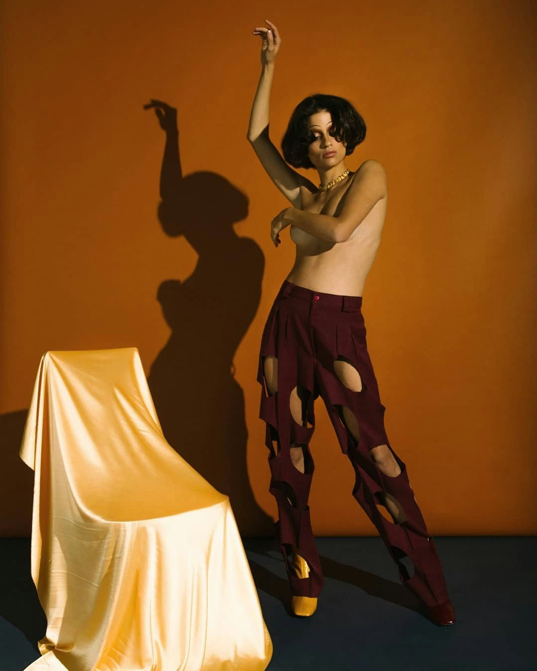 A young woman with stylish trousers and gold heels striking a pose against an orange backdrop with her shadow cast artistically.